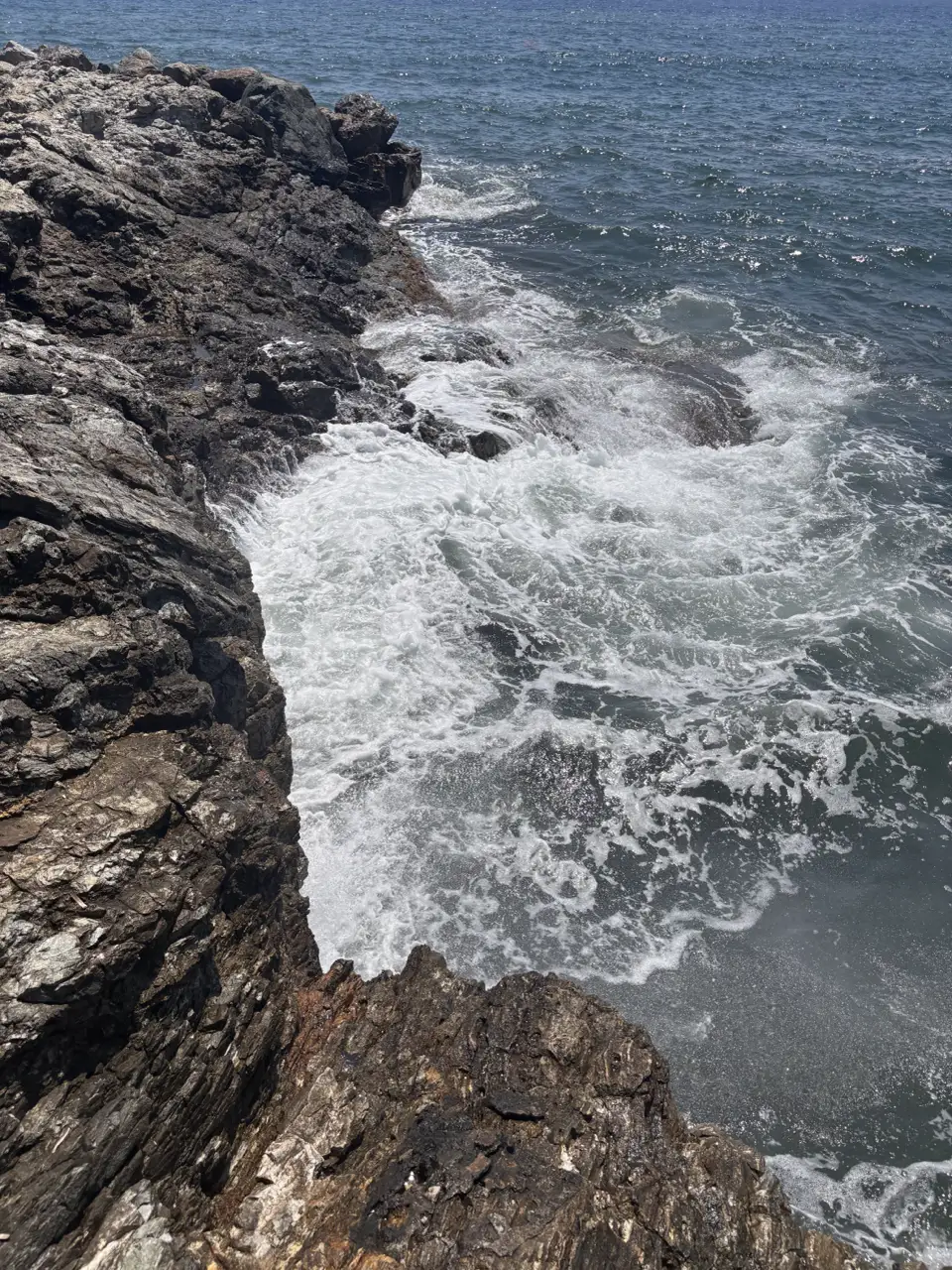 Brandung an der Felsküste: weiße Gischt bricht über dunkle Felsen, das Wasser dahinter ist aufgewühlt.