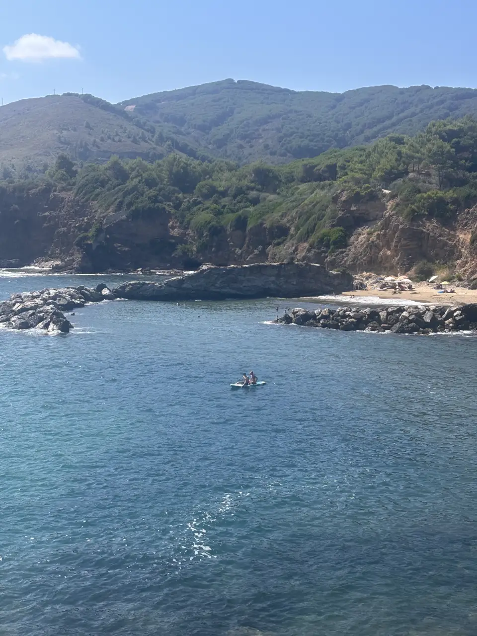 Blick von oben in die Bucht: ein heller Strandbogen, davor ein Felsriegel, der ein ruhiges Becken abtrennt; dahinter steigt ein bewaldeter Hang an.