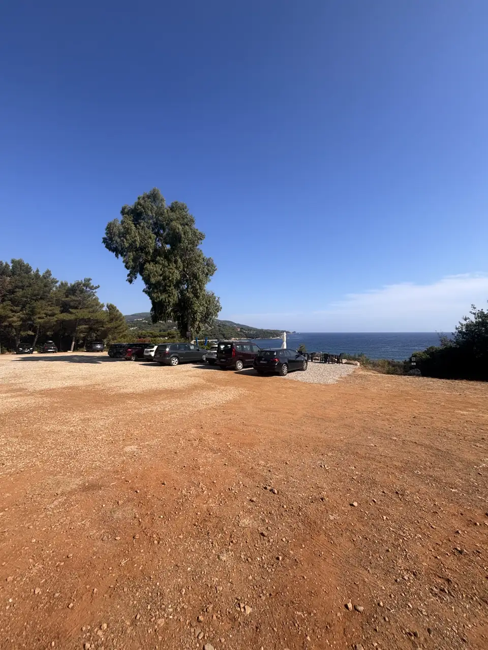 Der Parkplatz: eine offene Fläche aus rotem Erdreich, am Rand parkende Autos, dahinter Pinien und das Meer.