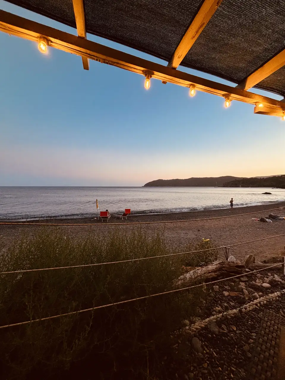 Blick aus dem überdachten Strandlokal über den Kiesstrand aufs Meer in der Abenddämmerung; unter dem Dach hängt eine Lichterkette, am Wasser stehen rote Liegestühle.