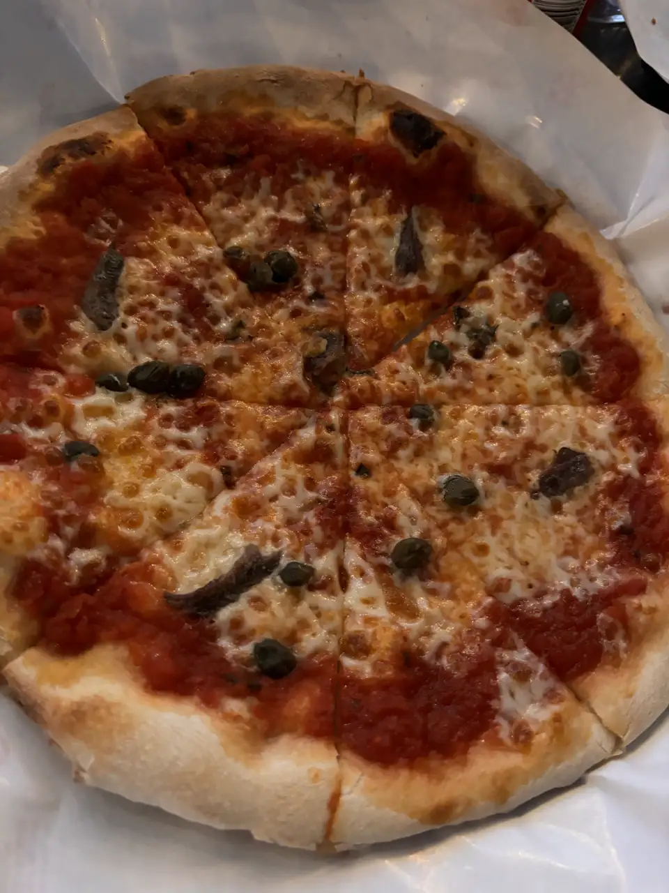 Eine Pizza mit Tomate, Käse und Oliven auf Backpapier, daneben eine zweite auf einem Metallteller.