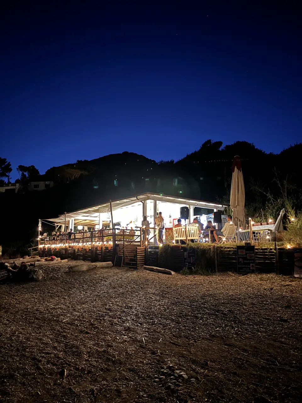 Das Strandlokal bei Nacht von außen: ein offener Pavillon mit hellem Licht und Lichterketten, davor Strand und dunkle Hügel.