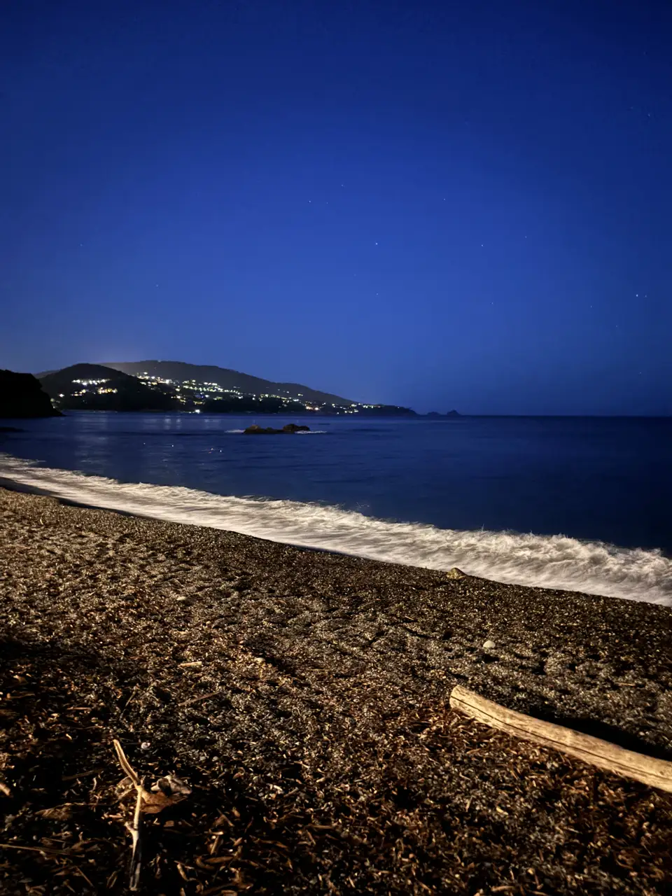 Der Strand bei Nacht: auslaufende Wellen auf dunklem Kies, am gegenüberliegenden Ufer die Lichter der Küste.