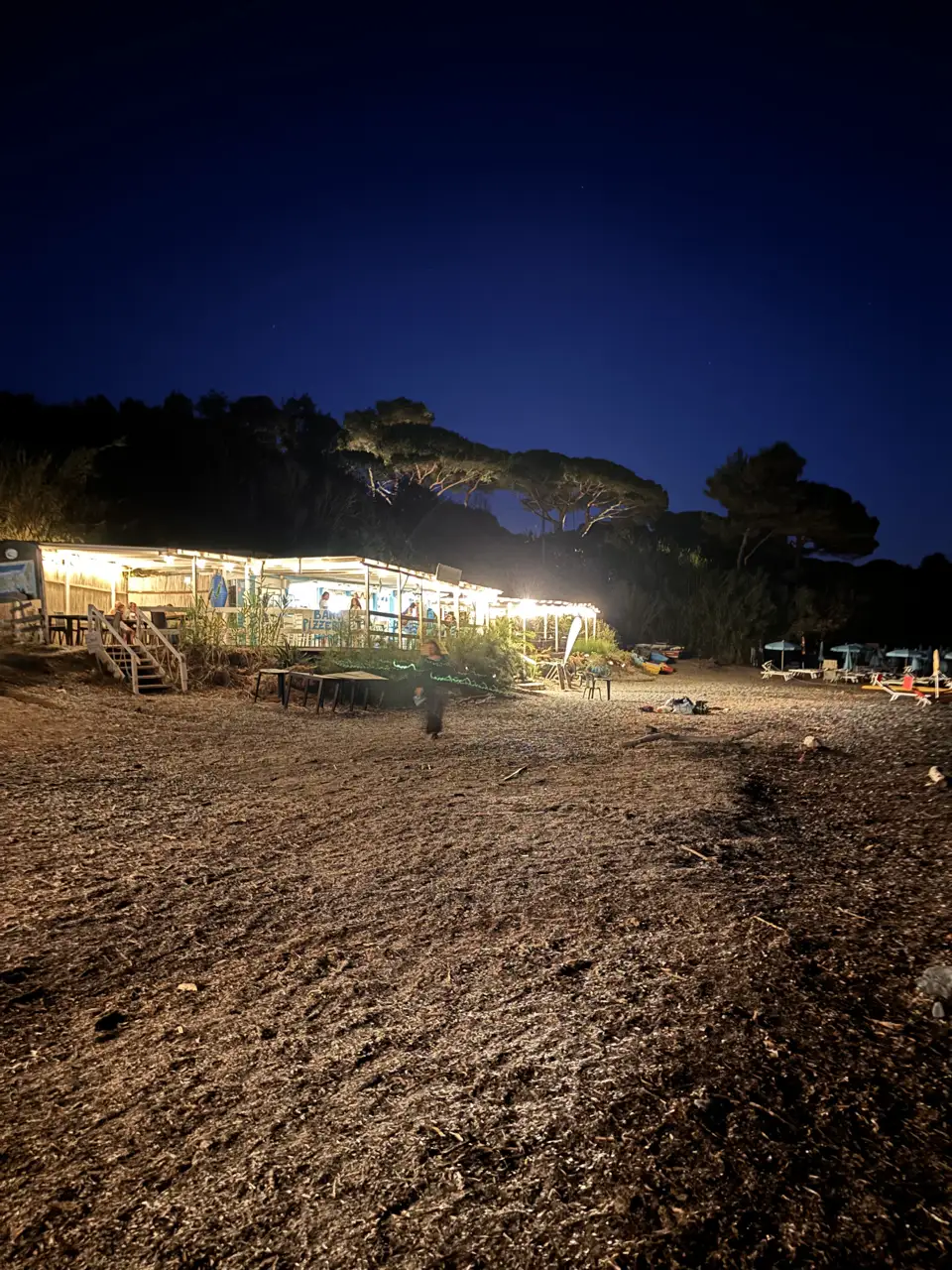 Das beleuchtete Lokal von der Seite, davor zusammengeklappte Liegen und Schirme auf dem Strand.