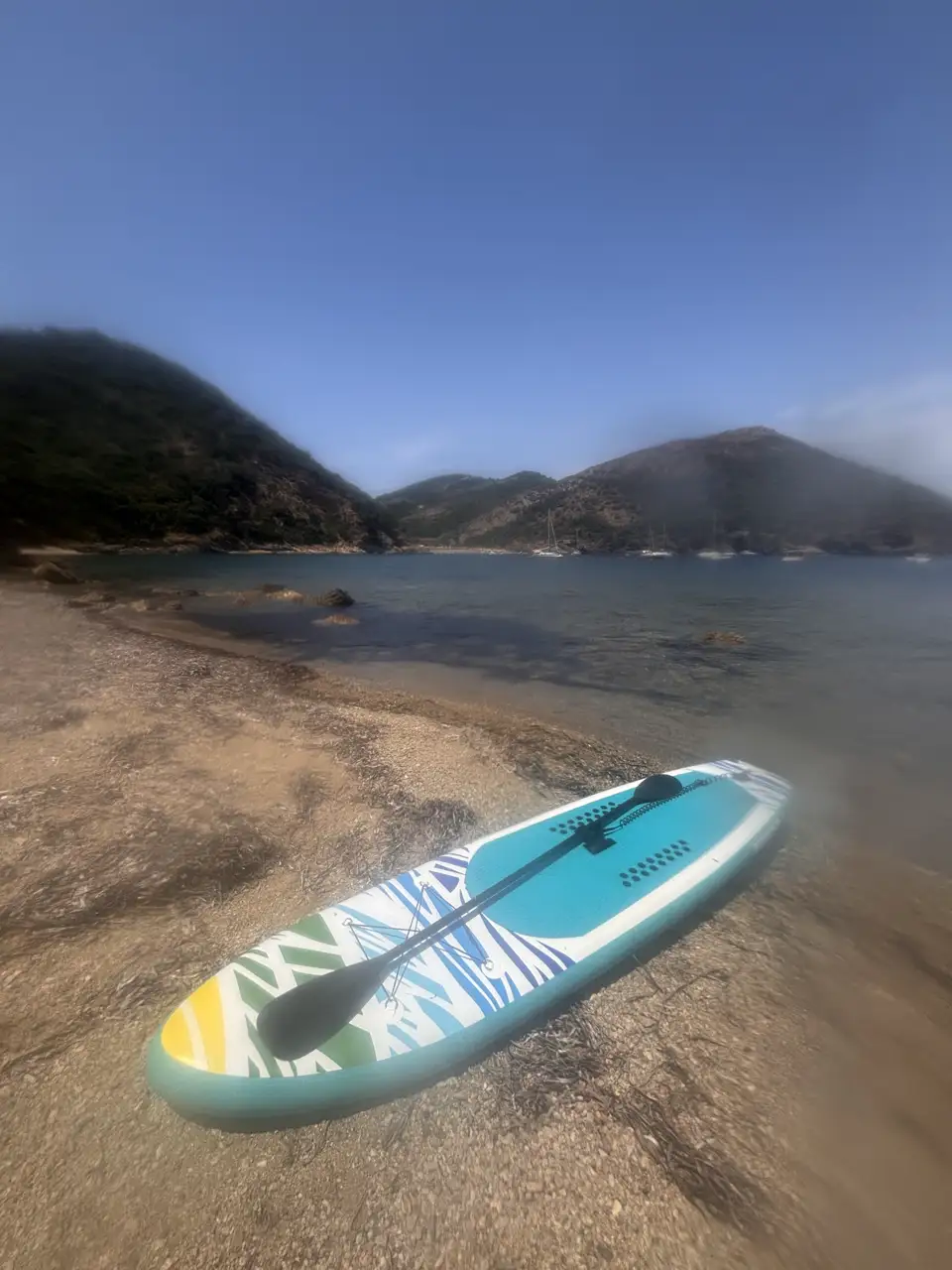 Ein Stand-up-Paddle-Board liegt an einem kleinen Kiesstrand zwischen Felsen.