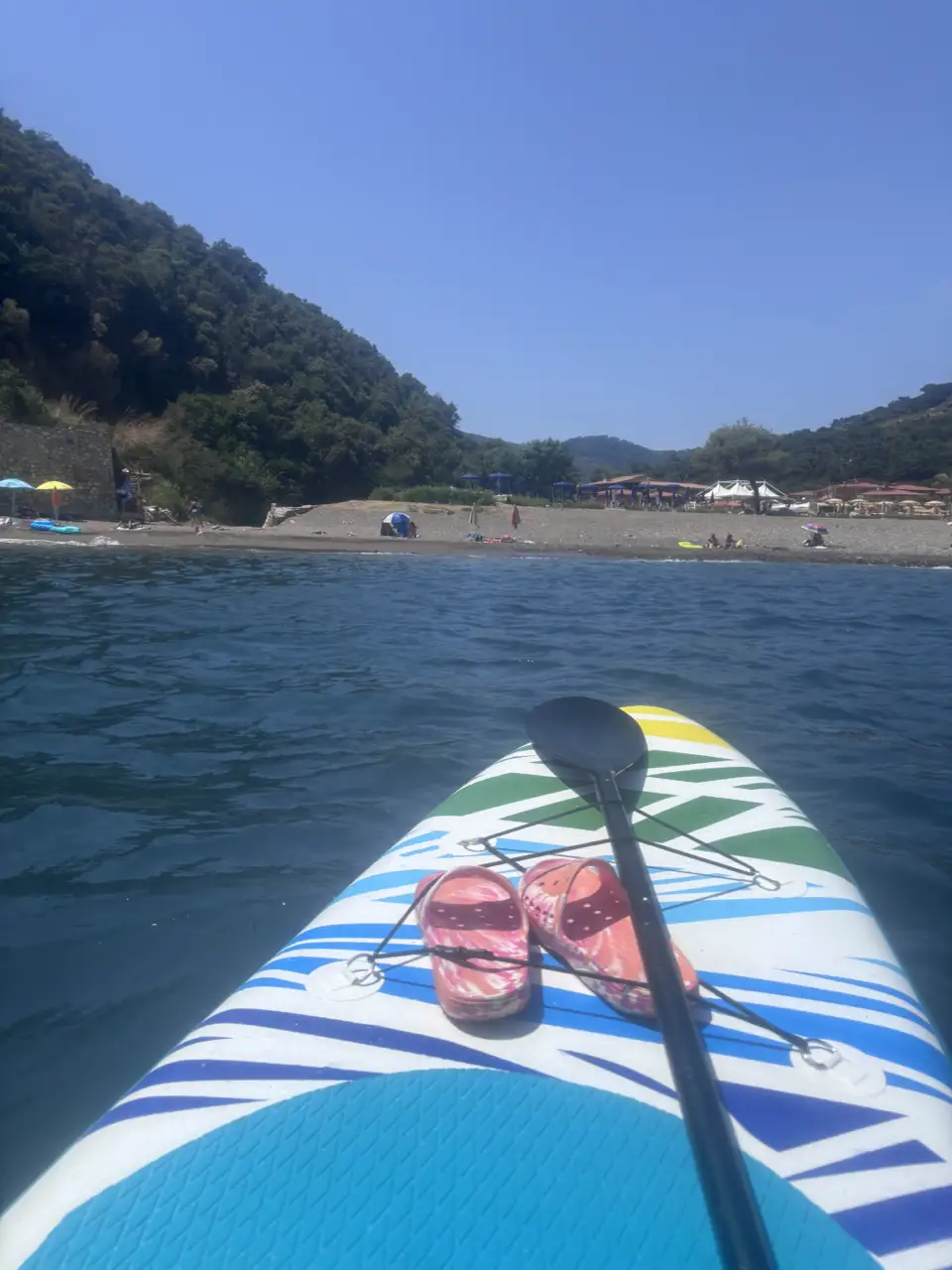 Vom Stand-up-Paddle-Board aus zurück auf den Strand: bewaldete Hänge rahmen die Bucht, am Ufer stehen Schirme.