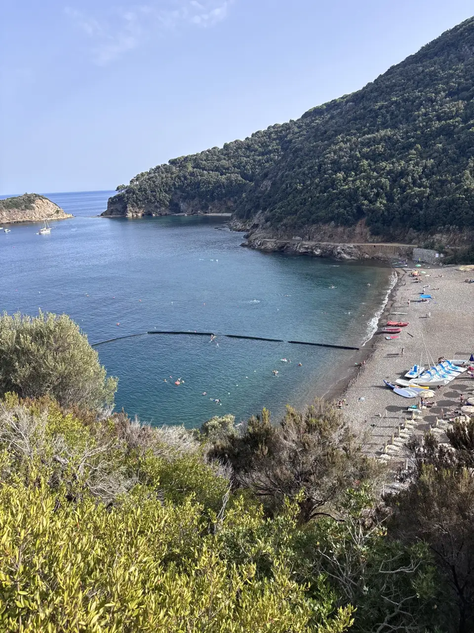 Die Bucht von oben: ein dunkler Kiesstrand im Bogen, davor eine Schwimmleine, am Ufer Liegen, Schirme und Tretboote. Links schließt eine bewaldete Landzunge die Bucht.