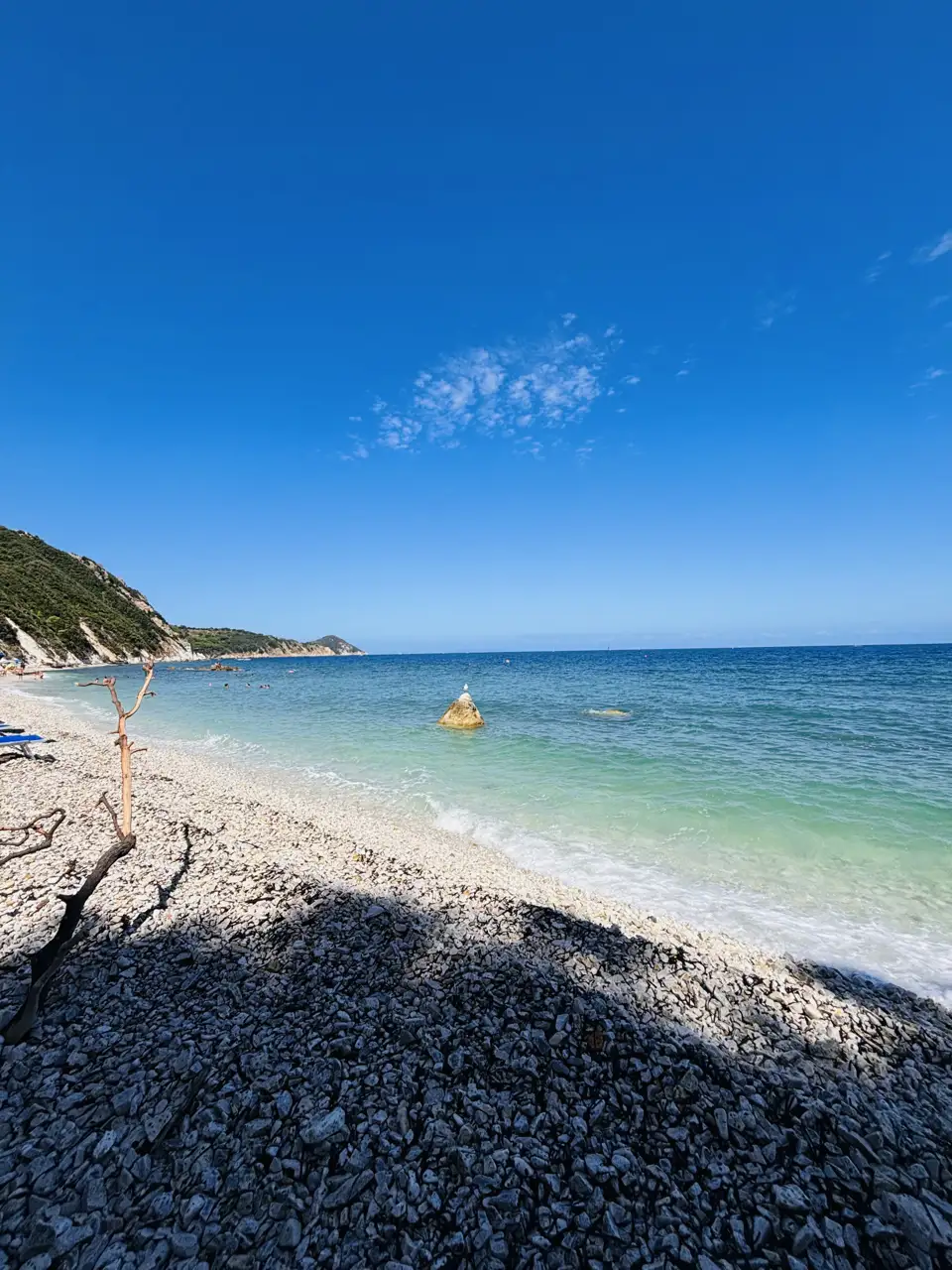Die Bucht von der Wasserlinie: heller, fast weißer Kiesstrand, davor türkis schimmerndes Wasser, im Hintergrund die bewaldete Landzunge.