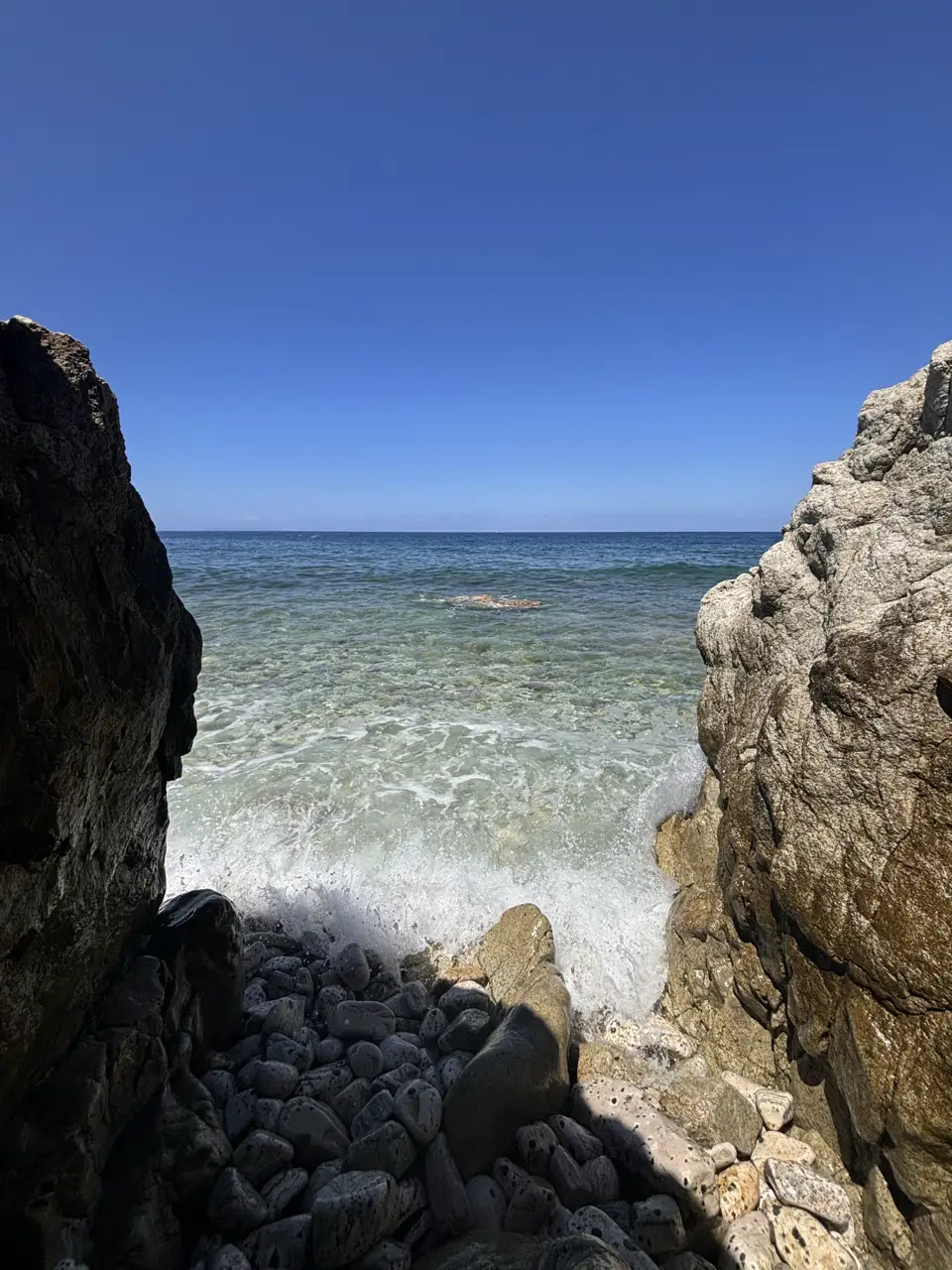 Zwischen zwei Felsblöcken hindurch der Blick auf klares Wasser; im Vordergrund runde helle Kiesel.