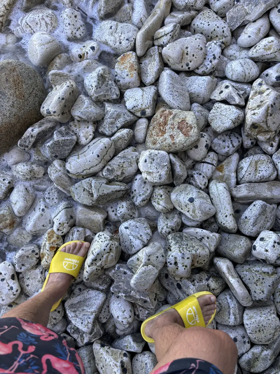 Nahaufnahme des Untergrunds: grober, weißer Kies, dazwischen einzelne rostrote Steine; am unteren Bildrand Badeschuhe.