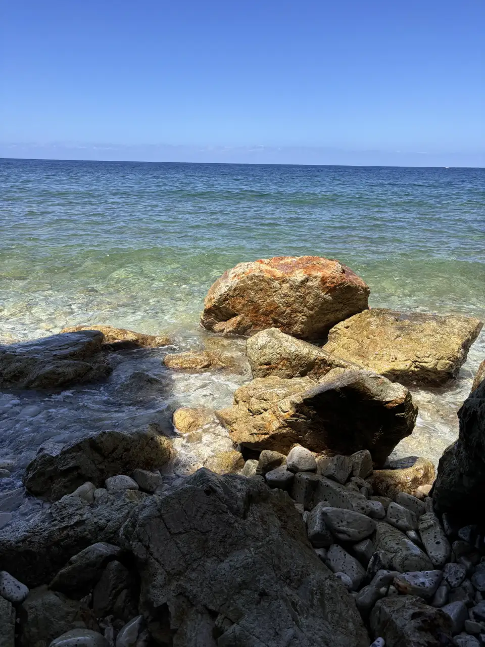 Rostrote und helle Felsblöcke am Ufer, dahinter ruhiges, klares Wasser bis zum Horizont.